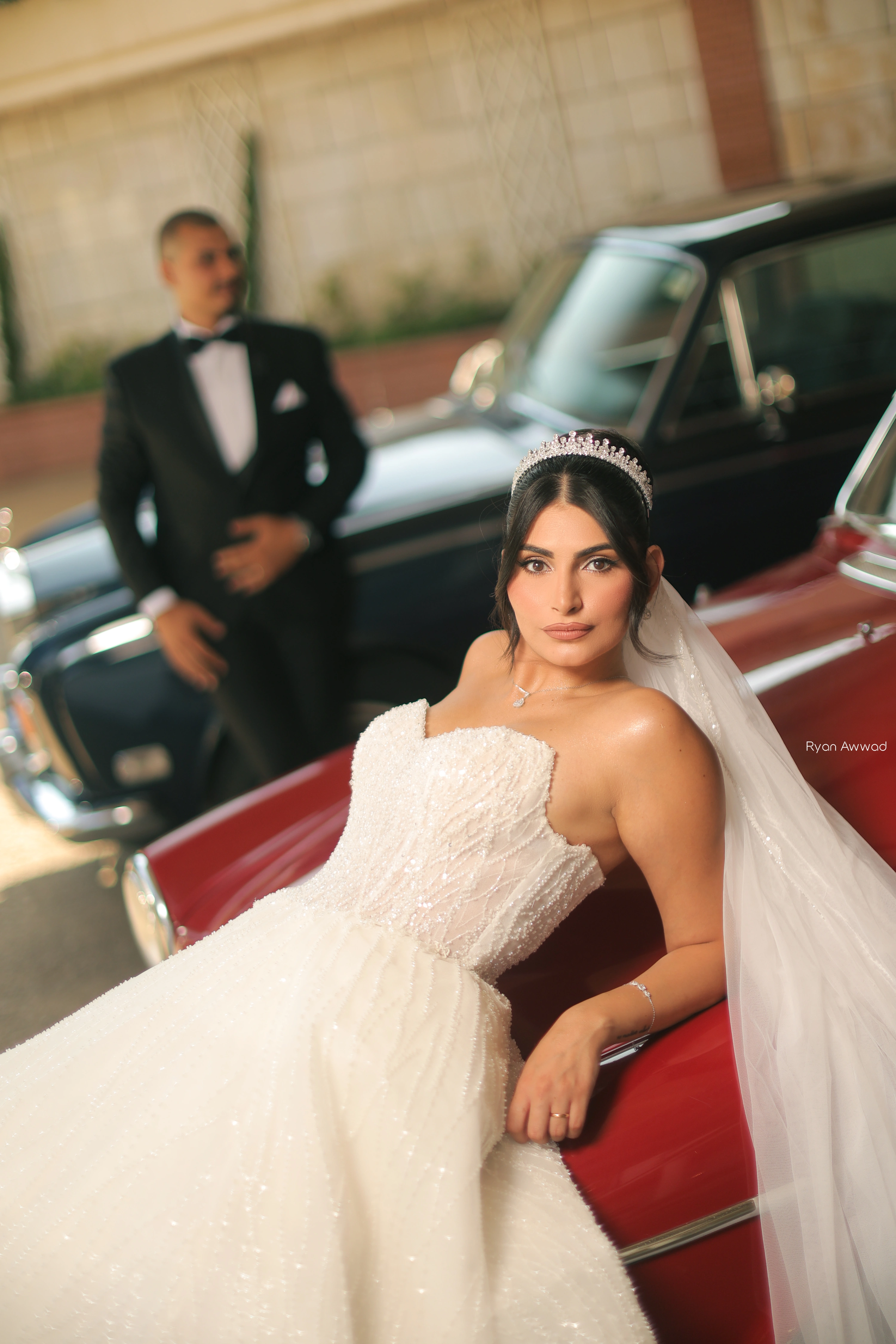 Bride seated on a red vintage car with the groom behind her